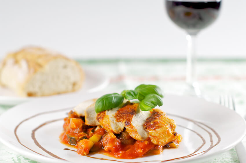 Eine Portion Hähnchenfilet mit roter Sauce und ein gefülltes Rotweinglas warten auf den Gast