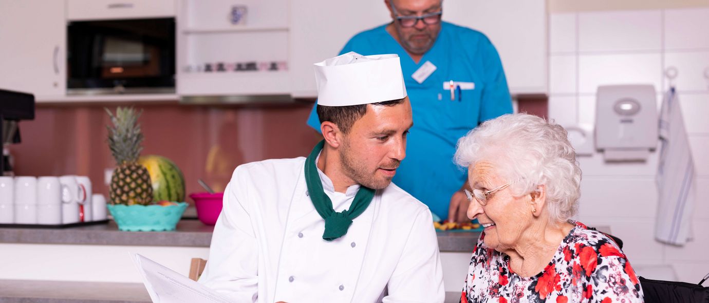 Koch mit Kochmütze serviert Seniorin Essen